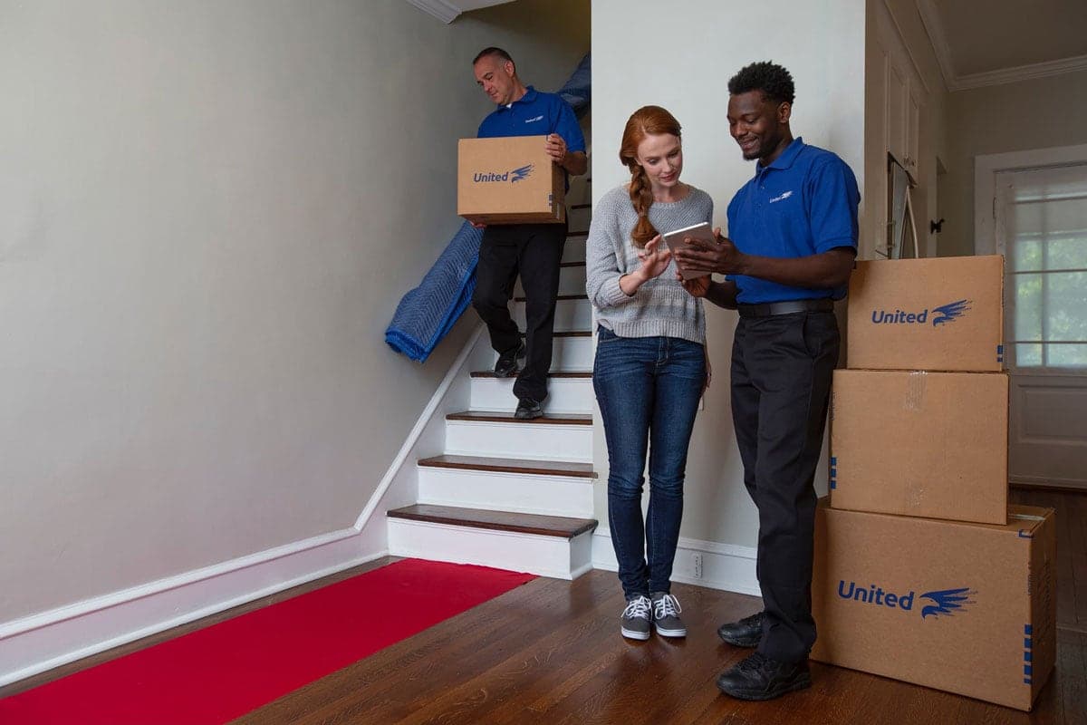 woman checks the shipping manifest with united mover while second mover takes moving boxes downstairs.